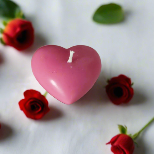 Bougie parfumée en forme de cœur rose entourée de roses rouges sur une surface blanche.