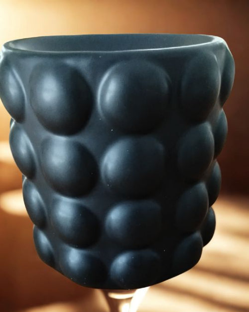Brûleur Bubble noir pour Fondants parfumés en céramique noire mate avec surface à reliefs ronds, posé sur une table en bois.