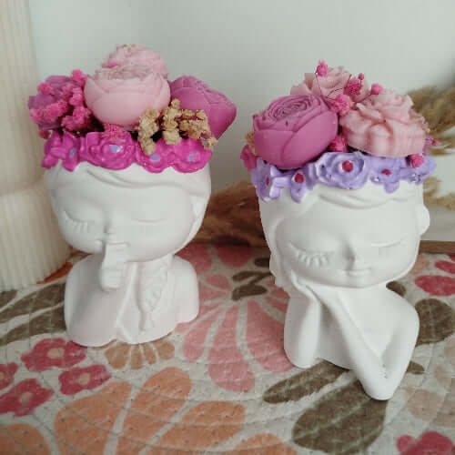 Deux bougies sculptées sur des supports en forme de visages féminins, décorées de fleurs en cire rose et violette.