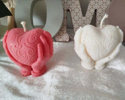 Bougie sculptée en forme de cœur rose tenue par des mains, posée sur une surface scintillante.
