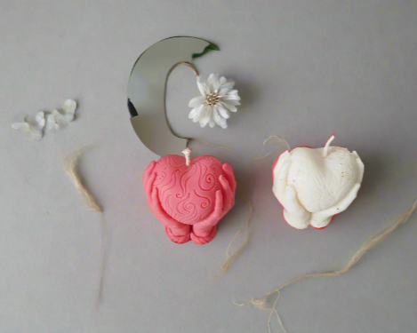 Deux bougies artisanales, l'une rose et l'autre blanche, en forme de cœur et mains, symboles de douceur et d'amour.
