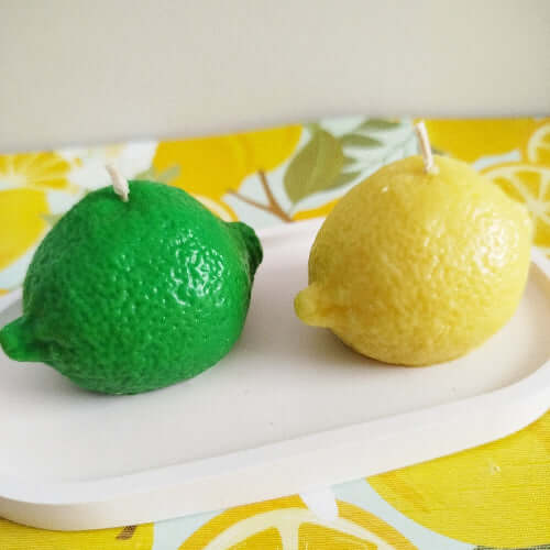 Deux bougies réalistes en forme de citron jaune et citron vert sur fond fruité.
